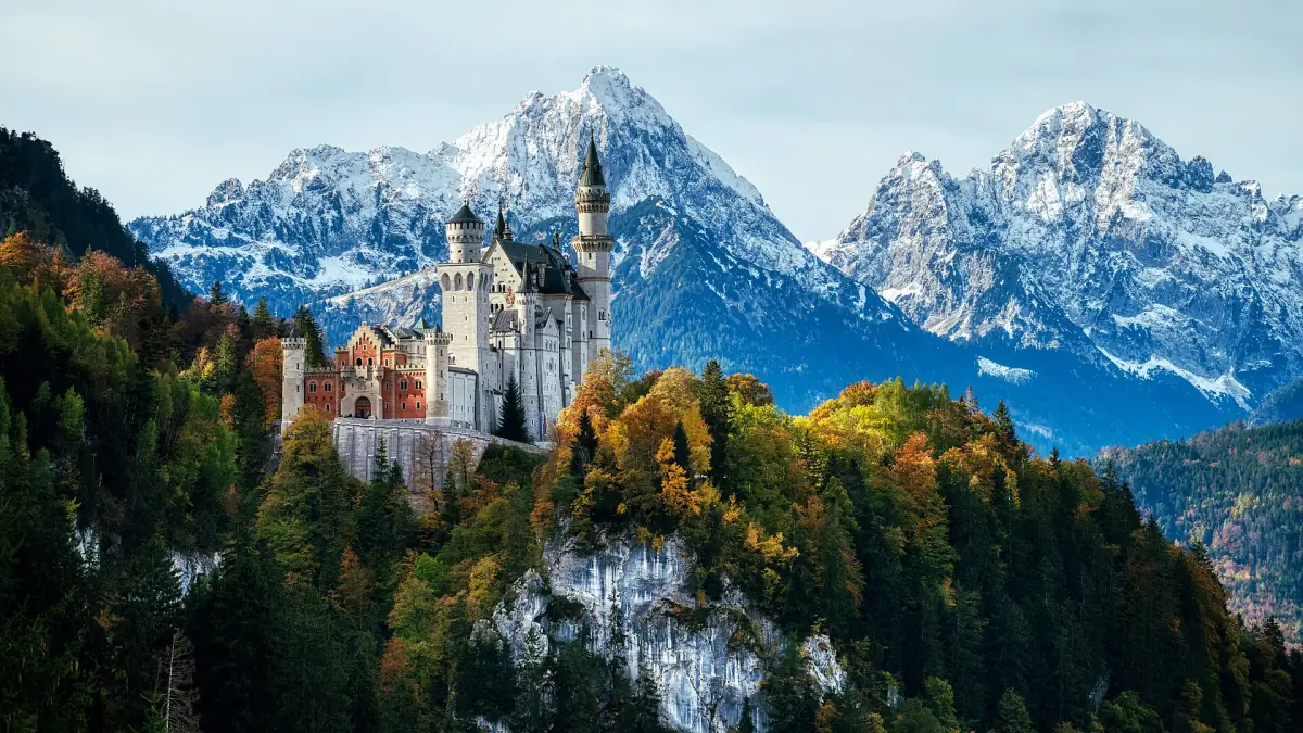 Neuschwanstein Castle above autumn forest with snow-capped peaks behind